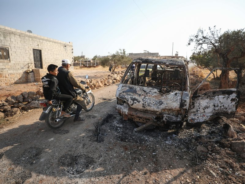 Syrians ride a motorcycle past a burned vehicle near the village of Barisha in the Idlib province of Syria, near where U.S. forces raided the compound of ISIS leader Abu Bakr al-Baghdadi. Local residents tell NPR that noncombatant civilians were killed and injured in the van the night of the U.S. raid. (via NPR from Omar Haj Kadour/AFP via Getty Images)