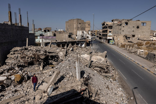 Yousef Hashim Ali, pictured here standing on the rubble of the building where 6 members of his family were killed. (Image from the New York Times)